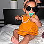 Zaria is registered to the contest to win money with this photo: toddler, child, pacifier, sunglasses, orange_dress, headband, bed, blanket, indoor, person, cute, fashion, accessory, hair, skin, foot, hand, furniture, home, portrait