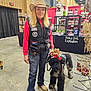 Nikita is registered to the contest to win money with this photo: woman, dog, cowboy_hat, vest, jeans, boots, indoor, market, holiday_decorations, reindeer_antlers, black_dog, crafts, smiling, leash, display_shelves, concrete_floor, casual_clothing, blonde_hair, shopping, festive