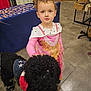 child, dog, indoor, curly_hair, dress, pink, black_dog, pet, playful, leash, concrete_floor, display_table, curious, girl, animal, friendship, costume, interaction, portrait, event