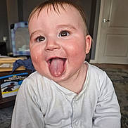 Theo joined the competition — help win amazing prizes! baby, child, tongue_out, white_clothing, onesie, happy, face, head, indoor, room, clock, carpet, furniture, hand, skin, tongue, person, smile, eyes, hair