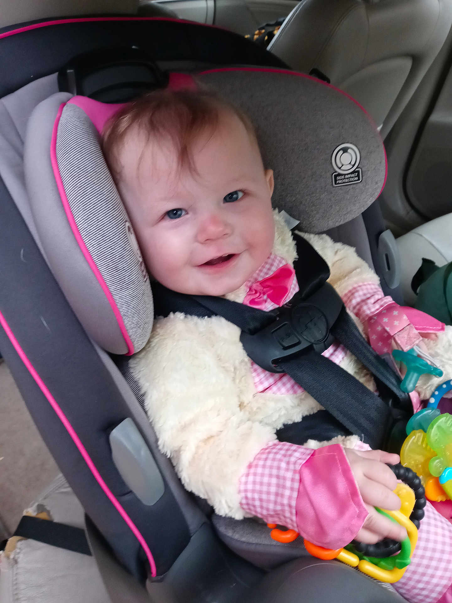 Allison is registered to the contest to win money with this photo: baby, car_seat, child, toy, pink_clothing, seat_belt, smiling, indoor, car_interior, blue_eyes, plush_jacket, hand, face, person, infant, safety_harness, cute, seat, child_safety, happy
