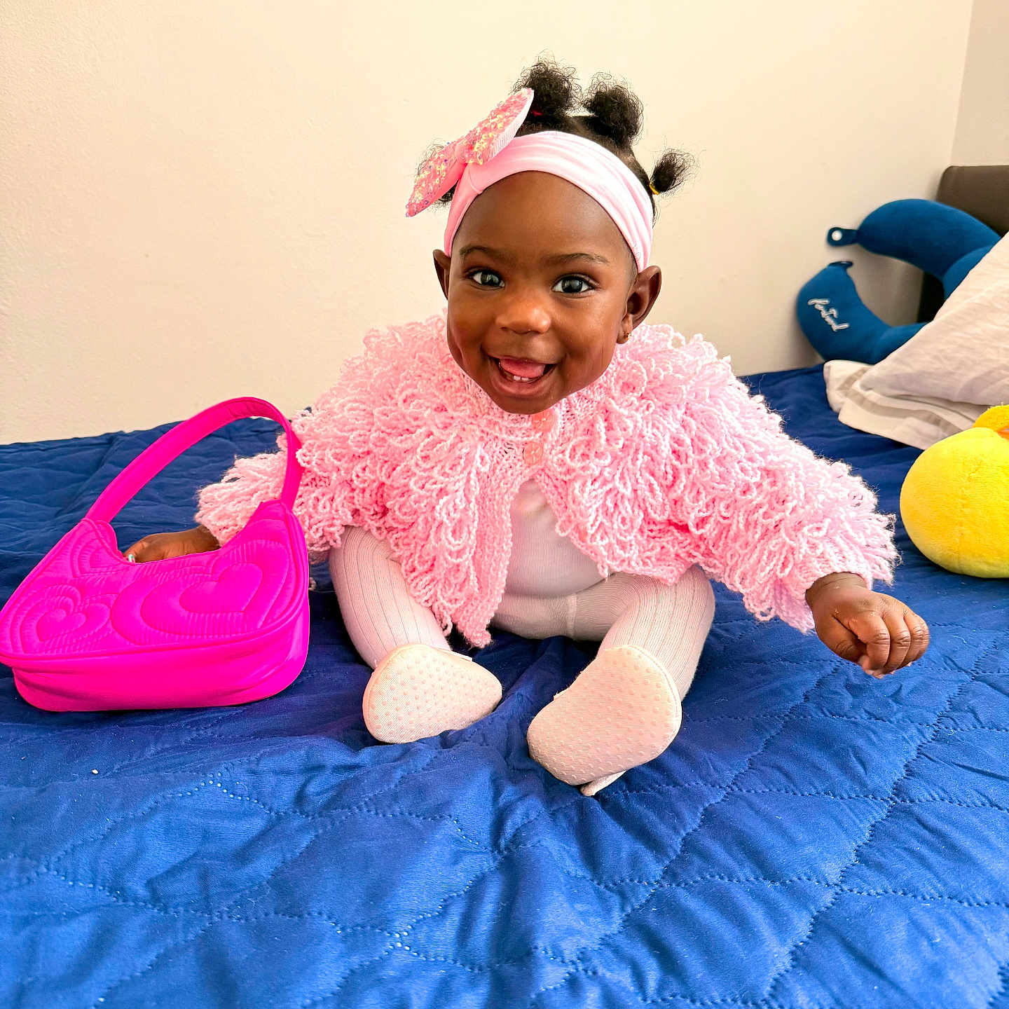Joanne is registered to the contest to win money with this photo: baby, blue_bedspread, child, clothing, colorful, cozy, cute, happy, headband, home, indoors, infant, person, pillow, pink_sweater, playful, portrait, purse, sitting, smiling