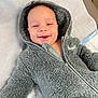 Elias is registered to the contest to win money with this photo: baby, smiling, hood, jacket, gray, fluffy, infant, child, happy, cute, warm, clothing, portrait, cozy, lying_down, indoor, person, face, young, soft