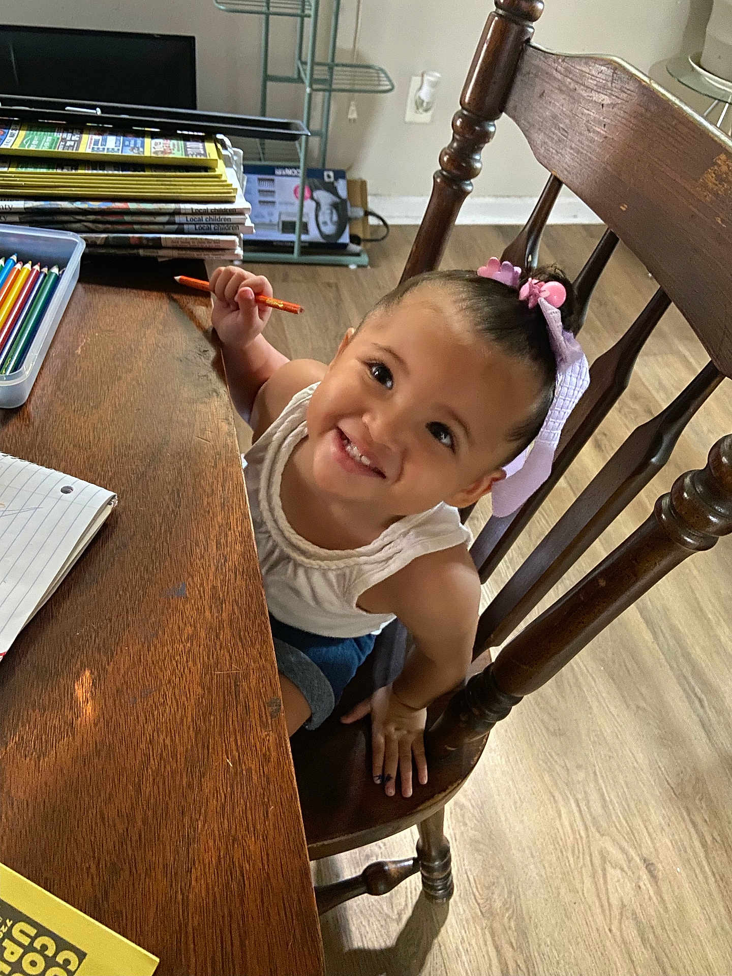 Analaya is registered to the contest to win money with this photo: child, toddler, smiling, colored_pencil, table, chair, wooden_furniture, hair_bow, white_shirt, notebook, floor, indoor, happy, person, writing, desk, art_supplies, cute, young_child, playful