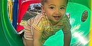 Analaya is registered to the contest to win money with this photo: toddler, child, playground, slide, green_tunnel, outdoor, sunlight, crawling, smiling, happy, park, plastic, colorful, young_child, casual_clothing, shorts, summer, daytime, nature, trees