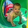 toddler, child, playground, slide, green_tunnel, outdoor, sunlight, crawling, smiling, happy, park, plastic, colorful, young_child, casual_clothing, shorts, summer, daytime, nature, trees