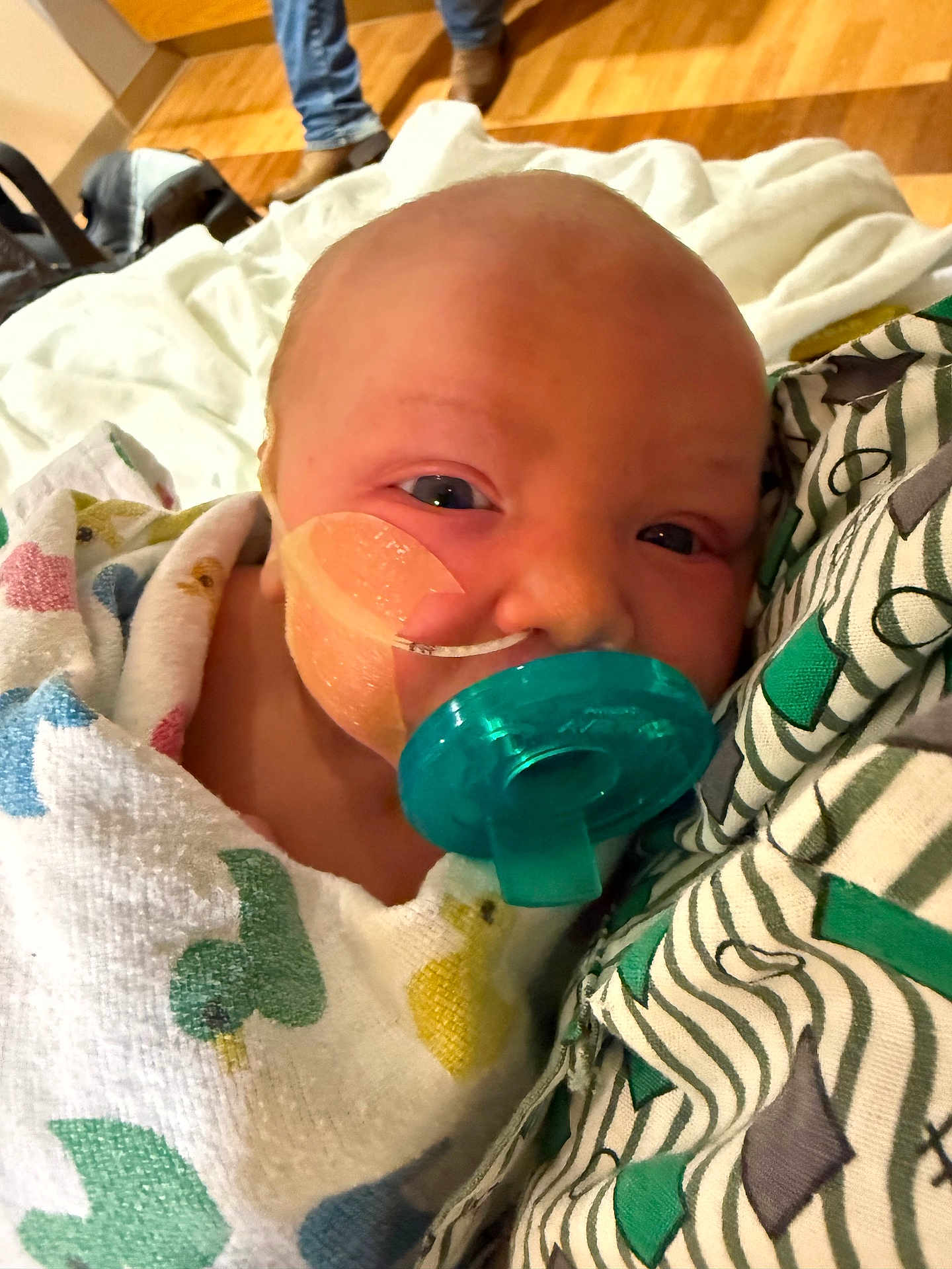 Isabella is registered to the contest to win money with this photo: baby, newborn, pacifier, blanket, medical_tape, face, infant, close_up, person, resting, indoor, floor, legs, clothing, patterned_fabric, comfort, child, swaddled, skin, head