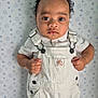 baby, child, overalls, white_clothing, curly_hair, bracelet, portrait, indoor, star_pattern, background, cute, young_child, skin, face, eyes, hands, fashion, infant, standing, expression