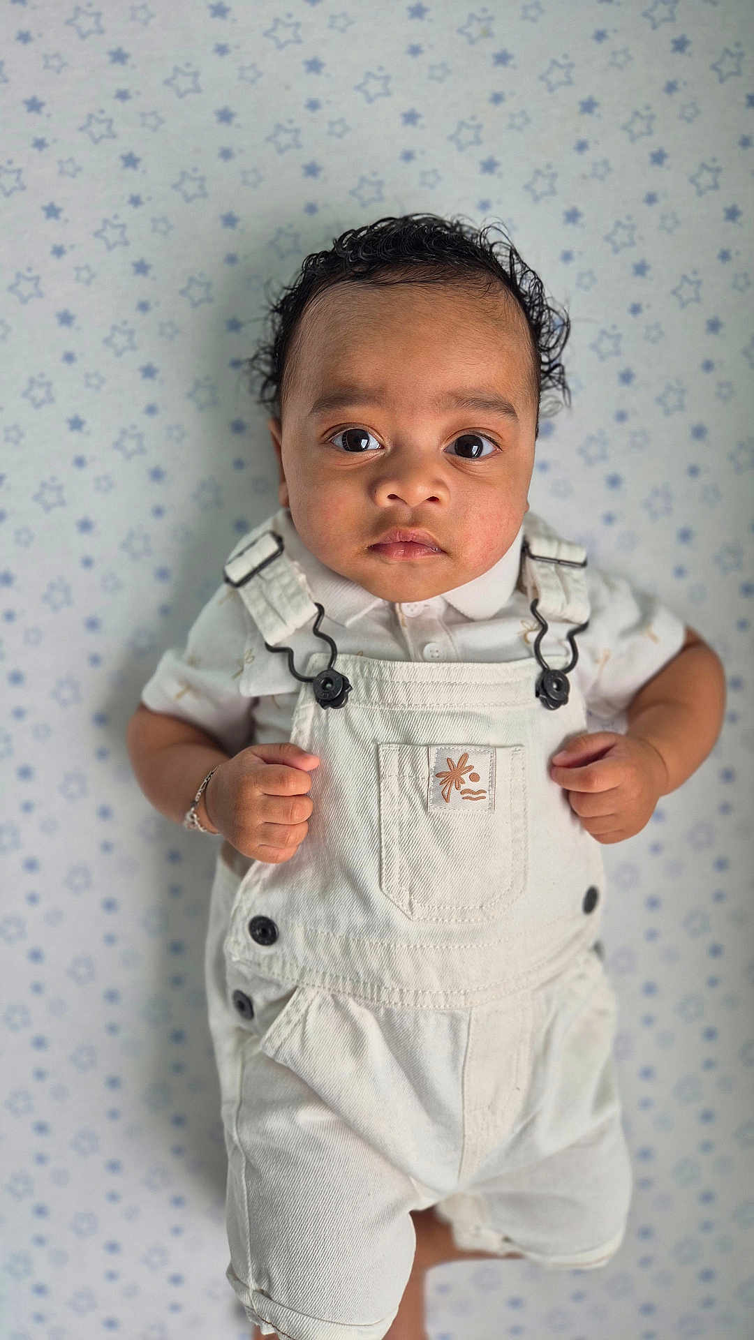 Aiden participe au concours pour gagner de l'argent avec cette photo : baby, child, overalls, white_clothing, curly_hair, bracelet, portrait, indoor, star_pattern, background, cute, young_child, skin, face, eyes, hands, fashion, infant, standing, expression