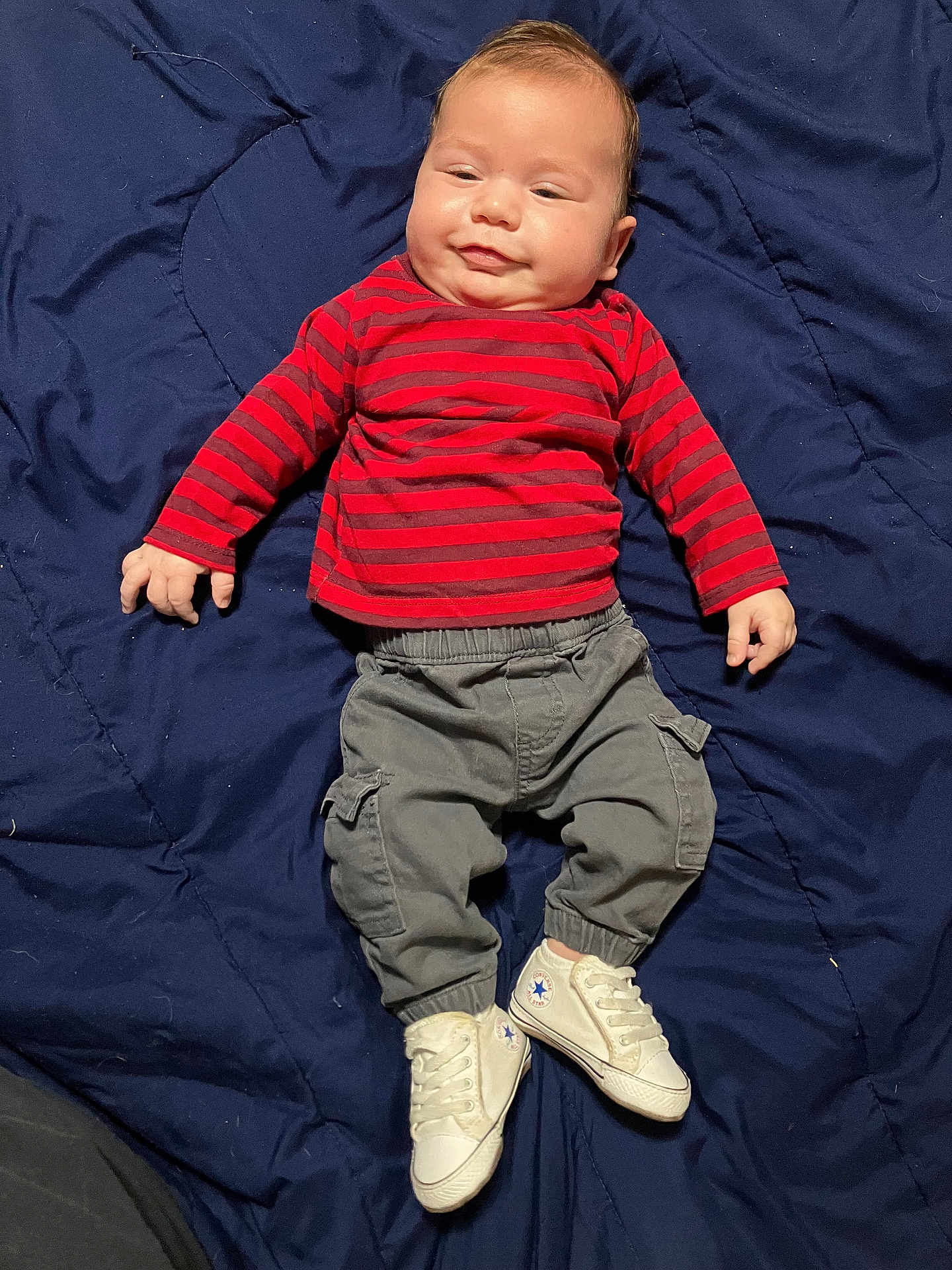 Raiden is registered to the contest to win money with this photo: baby, infant, child, striped_shirt, red, gray_pants, cargo_pants, white_sneakers, footwear, blanket, blue_blanket, laying_down, smiling, face, hands, cute, indoors, casual_clothing, small_feet, young