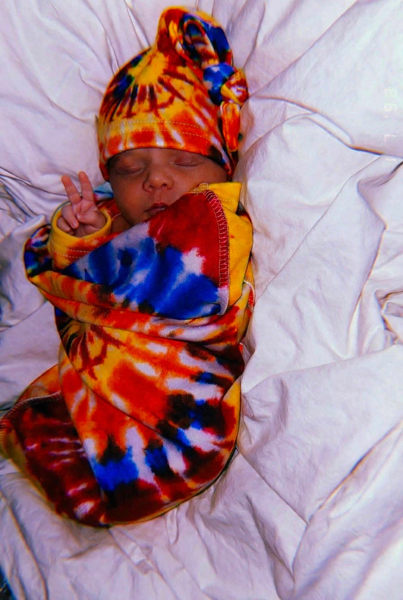 Sophia is registered to the contest to win money with this photo: baby, sleeping, peace_sign, tie_dye, blanket, hat, colorful, infant, bed, bedding, cute, peaceful, wrapped, soft, newborn, child, portrait, cozy, rest, warm