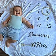 Santino participe au concours pour gagner de l'argent avec cette photo : baby, infant, smiling, milestone_blanket, numbers, wooden_ring, blue_clothing, light_blue, shorts, sleeveless_top, happy, lying_down, white_blanket, pom_pom_edge, celebration, french_text, semaines, mois, floor, person