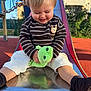 toddler, child, playground, slide, plush_toy, green_frog, smile, striped_shirt, white_pants, socks, hands, legs, outdoor, park, candid, portrait, metal_slide, sunlight, joy, parent_hand