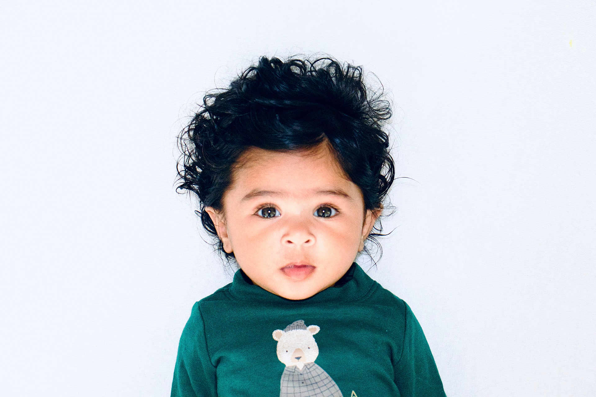 Swaha joined the competition — help win amazing prizes! toddler, child, curly_hair, green_shirt, bear_illustration, portrait, face, eyes, cute, indoor, white_background, young_child, expression, clothing, baby, person, headshot, looking_at_camera, studio_photo, innocence