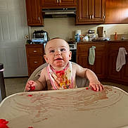 Molly is registered to the contest to win money with this photo: appliance, baby, bib, cabinet, child, countertop, face, food, hand, happy, high_chair, indoor, kitchen, messy, person, refrigerator, sink, smile, table, towel