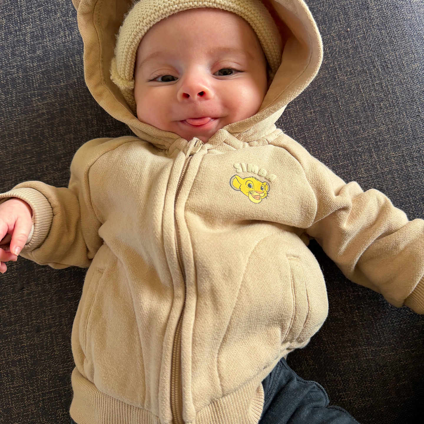 Tiago participe au concours pour gagner de l'argent avec cette photo : baby, child, hoodie, beige, smile, tongue_out, knitted_hat, cute, playful, person, clothing, fabric, dark_background, portrait, infant, sitting, warm, casual, face, indoors