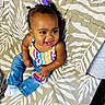 child, baby, toddler, portrait, smiling, sitting, bed, bedspread, patterned_background, rainbow_top, striped_clothing, jeans, bow, hair_accessory, earrings, socks, indoor, happy, fashion, close_up