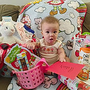 Antania is registered to the contest to win money with this photo: baby, infant, onesie, blanket, couch, plush_toy, basket, toy_packaging, card, surprised_expression, sitting, gift, pink, strawberry, snack_pouch, decorative_blanket, home_interior, hand, eyes, portrait
