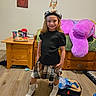 backpack, bedroom, black_top, boots, child, cups, dreamcatcher, flooring, girl, hat, indoor, keyboard, plaid_skirt, posing, purple_plush, smile, snacks, spiderman_poster, stuffed_animal, wooden_table