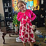 bottle, child, cup, dress, face, floor, furniture, girl, happy, indoor, living_room, pink_cardigan, plant, remote_control, shoes, smile, snacks, table, television, wooden_floor