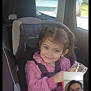 Scarlett is registered to the contest to win money with this photo: child, car_seat, pink_sweater, smiling, video_call, snack, vehicle_interior, seatbelt, girl, window, car, happy, person, glasses, face, technology, screen, communication, casual_clothing, daytime