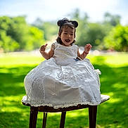 Sophia joined the competition — help win amazing prizes! child, toddler, smiling, white_dress, outdoor, greenery, grass, wooden_stool, happy, portrait, sunlight, nature, cute, sitting, hair_buns, daylight, young_child, celebration_dress, joyful, person