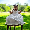 child, toddler, smiling, white_dress, outdoor, greenery, grass, wooden_stool, happy, portrait, sunlight, nature, cute, sitting, hair_buns, daylight, young_child, celebration_dress, joyful, person