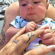 Braxtyn joined the competition — help win amazing prizes! adult, baby, background, closeup, clothing, curious, expression, face, fingers, furniture, hand, holding, indoor, infant, interaction, laundry, onesie, person, skin, tattoo