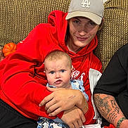 Adalee is registered to the contest to win money with this photo: baby, child, young_person, red_hoodie, gray_cap, blue_eyes, butterfly_pattern, pants, tattoo, arm, couch, seated, hand, expression, indoor, fabric, clothing, person, face, skin