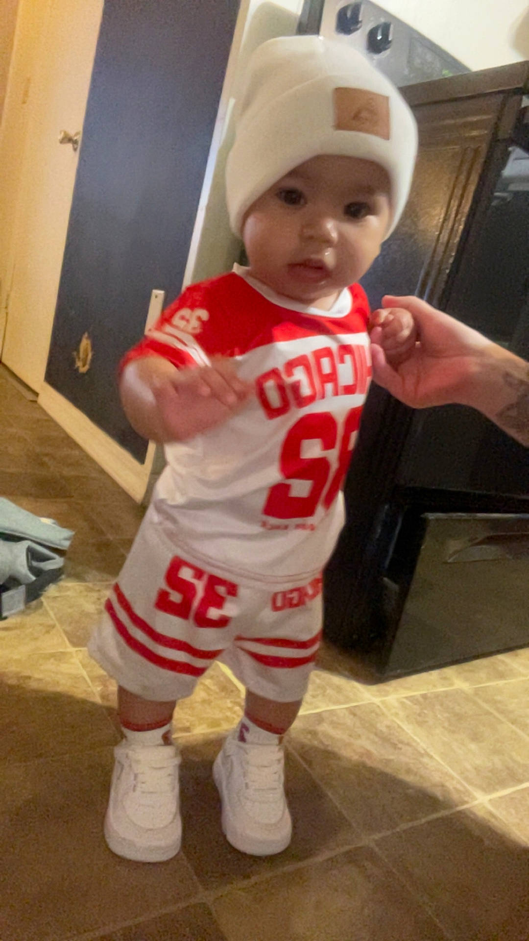 adult_hand, baby, beanie, child, cute, floor, full_body, furniture, home, indoor, jersey, portrait, red_clothing, sneakers, sporty_outfit, standing, television, tiles, toddler, white_clothing