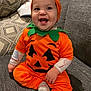 baby, child, pumpkin_costume, orange_clothing, smiling, happy, couch, gray_fabric, cushions, patterned_pillows, indoor, cute, holiday_costume, seated, infant, face, headwear, tooth, cheerful, portrait