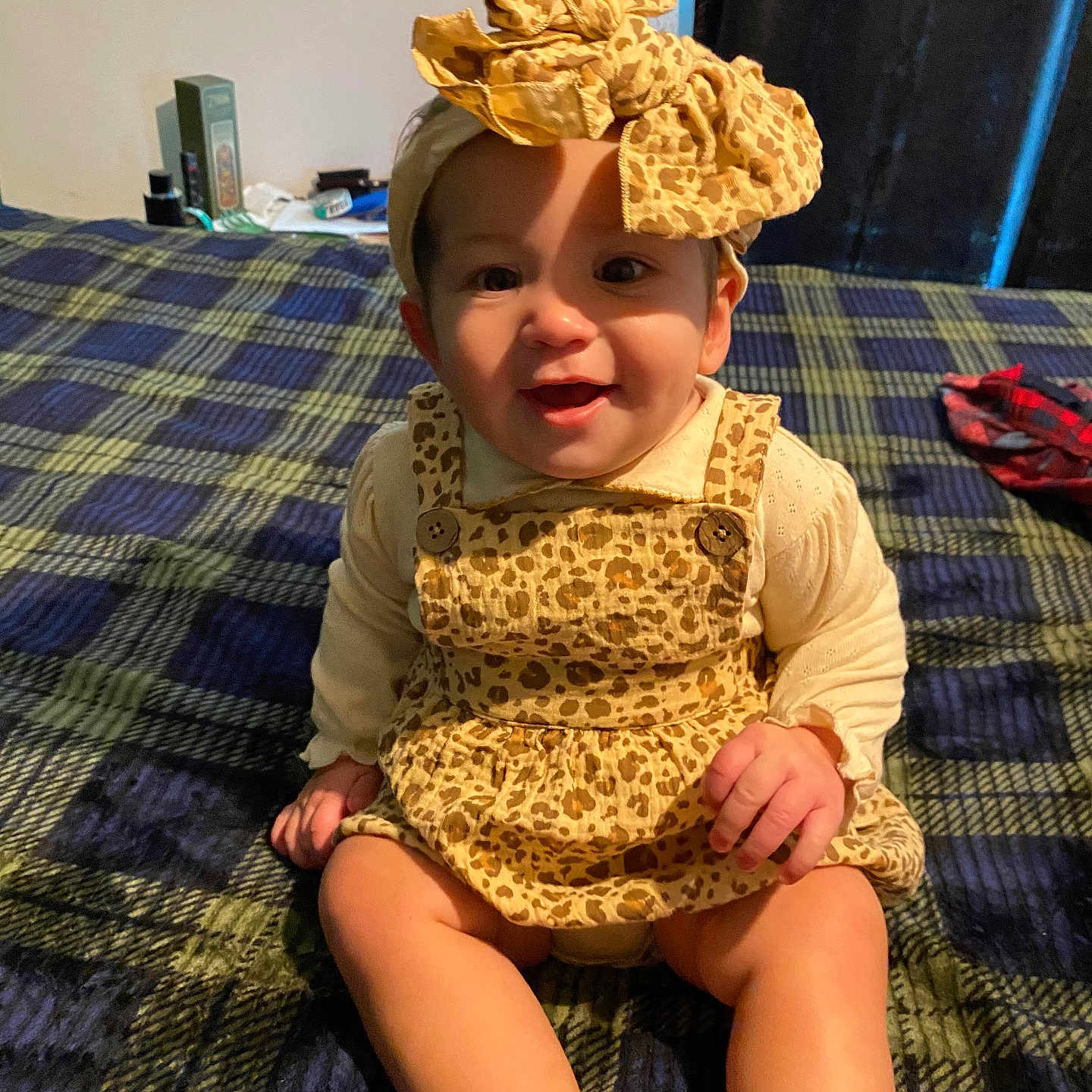 Arya is registered to the contest to win money with this photo: baby, child, clothing, cute, dress, face, hands, happy, headband, headwear, indoors, infant, legs, leopard_print, person, plaid_blanket, portrait, sitting, smiling, young