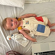 Roman participe au concours pour gagner de l'argent avec cette photo : toddler, child, bed, pillow, plush_toy, duck, overalls, laptop, red_bag, clothing, face, person, indoor, blanket, furniture, curious, baby, head, hand, foot