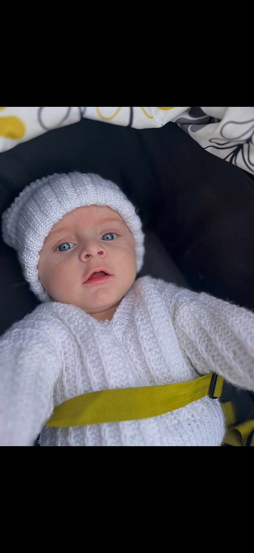 Ardonis joined the competition — help win amazing prizes! baby, child, blue_eyes, knitted_hat, knitted_sweater, white_clothing, yellow_harness, close_up, portrait, infant, cute, warm_clothing, car_seat, seatbelt, person, young_child, indoor, face, expression, soft_texture