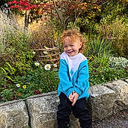 Enver joined the competition — help win amazing prizes! child, toddler, curly_hair, smiling, blue_sweater, black_pants, black_shoes, stone_wall, garden, flowers, greenery, daisies, autumn, outdoor, nature, plants, grass, tree, happy, portrait