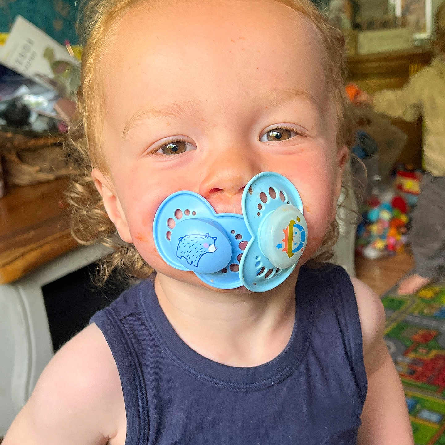 Enver is registered to the contest to win money with this photo: baby, blue_shirt, candid, child, childhood, curly_hair, cute, expression, face, funny, home, indoor, pacifier, person, playroom, portrait, smile, toddler, toys, young_child