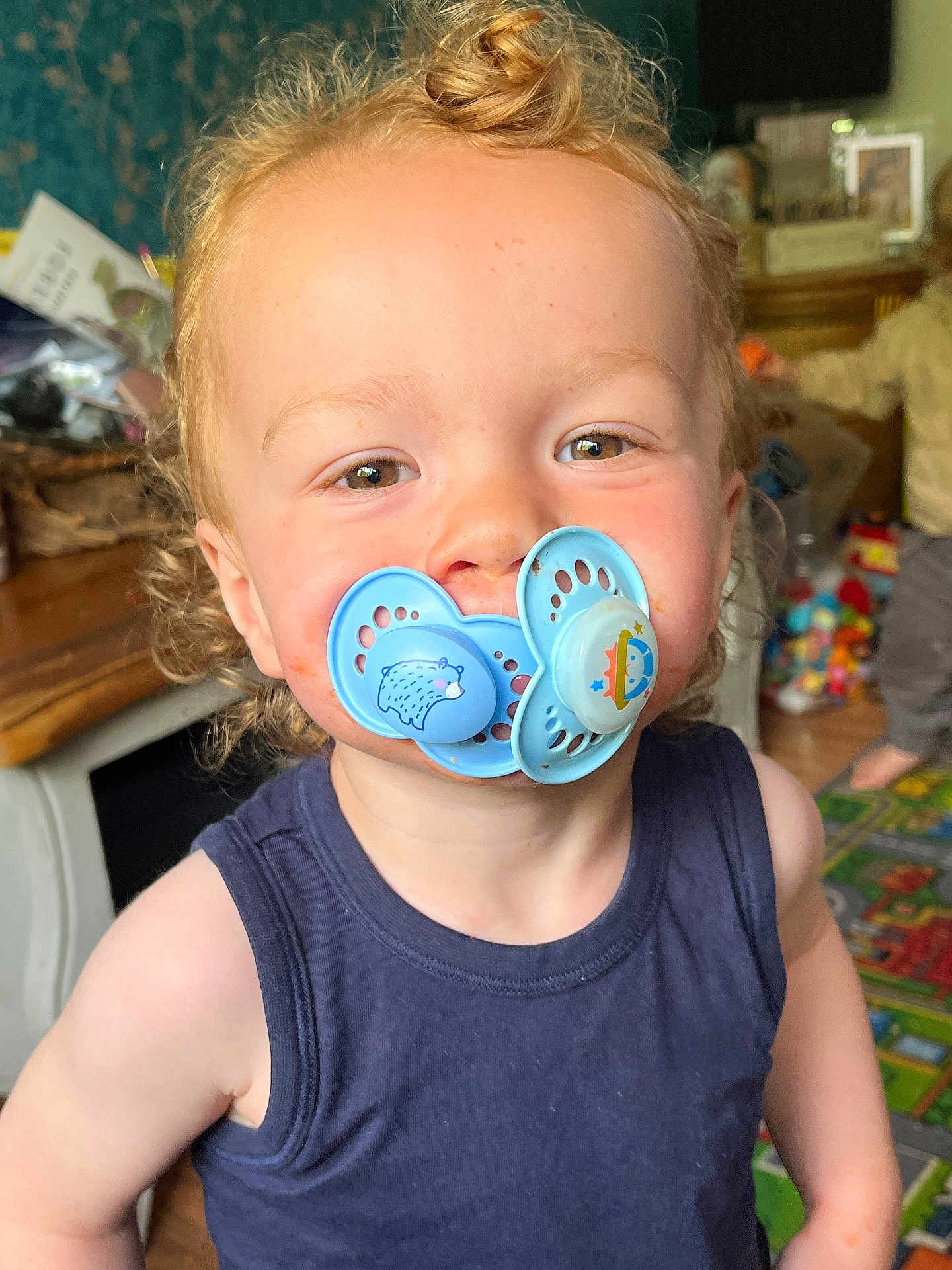 Enver is registered to the contest to win money with this photo: toddler, child, pacifier, curly_hair, indoor, playroom, toys, blue_shirt, face, portrait, smile, baby, person, young_child, home, candid, cute, expression, funny, childhood