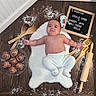 baby, baking_utensils, child, chubby, cinnamon_rolls, cooking, cute, expression, flour, food, indoors, infant, kitchen, letter_board, lying_down, measuring_spoons, rolling_pin, white_rug, wooden_floor, wooden_spoons