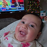 Taryn is registered to the contest to win money with this photo: baby, smiling, face, floral_clothing, indoors, christmas_tree, holiday_lights, television, news_program, couch, blanket, happy, child, portrait, infant, home, decor, bright_eyes, cute, joyful