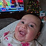 baby, smiling, face, floral_clothing, indoors, christmas_tree, holiday_lights, television, news_program, couch, blanket, happy, child, portrait, infant, home, decor, bright_eyes, cute, joyful