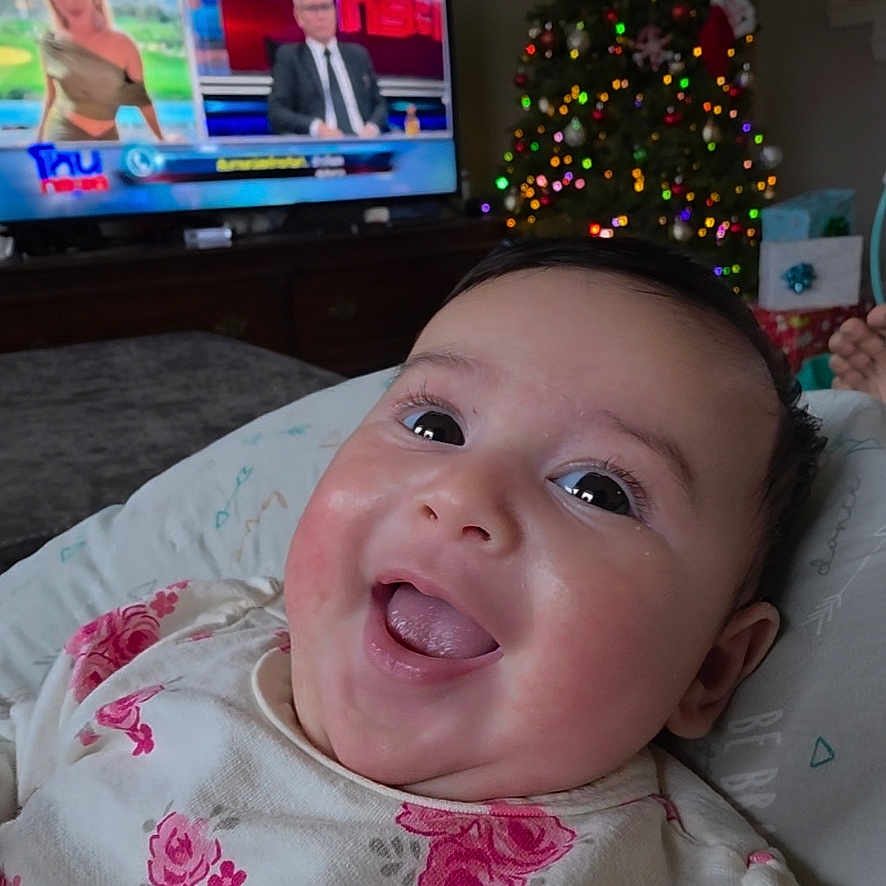 Taryn is registered to the contest to win money with this photo: baby, blanket, bright_eyes, child, christmas_tree, couch, cute, decor, face, floral_clothing, happy, holiday_lights, home, indoors, infant, joyful, news_program, portrait, smiling, television