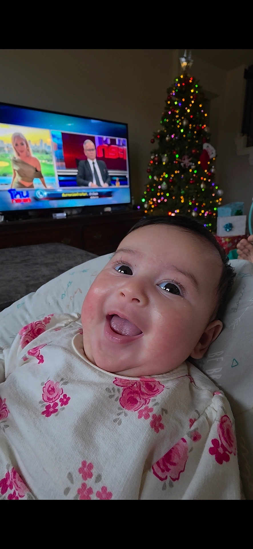 Taryn is registered to the contest to win money with this photo: baby, smiling, face, floral_clothing, indoors, christmas_tree, holiday_lights, television, news_program, couch, blanket, happy, child, portrait, infant, home, decor, bright_eyes, cute, joyful