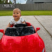 Kayden is registered to the contest to win money with this photo: baby, child, infant, smiling, happy, portrait, toy_car, red_car, sports_car, steering_wheel, outdoor, driveway, backyard, grass, fence, house, palm_tree, vehicle, milestone, text_overlay
