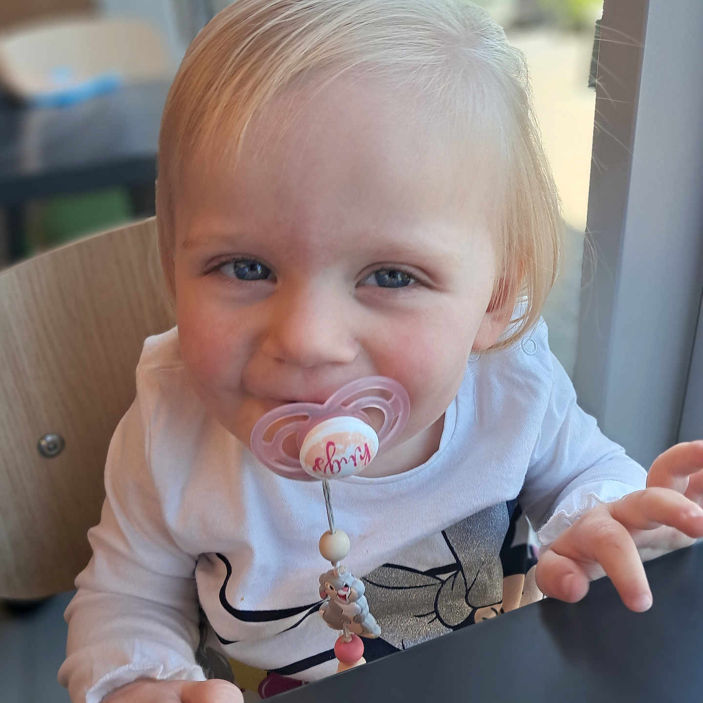 Mayla a rejoint le concours — aidez-le/la à gagner de superbes lots ! baby, blonde_hair, blue_eyes, chair, child, closeup, cute, face, hand, indoor, natural_light, pacifier, person, seated, smile, table, toddler, toy, white_shirt, window