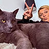 black_clothing, blonde_hair, cat, close_up, couch, eyes, face, funny, fur, gray_cat, home, indoor, lamp, peace_sign, person, pet, portrait, puckered_lips, whiskers, woman