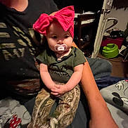Ameiire is registered to the contest to win money with this photo: baby, child, pacifier, headband, red_bow, person, sitting, indoor, clothing, camouflage_pants, arm, blanket, furniture, room, door, toy, carpet, casual, comfort, relaxation