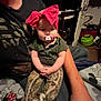 baby, child, pacifier, headband, red_bow, person, sitting, indoor, clothing, camouflage_pants, arm, blanket, furniture, room, door, toy, carpet, casual, comfort, relaxation