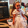Camila is registered to the contest to win money with this photo: child, girl, white_dress, lace, indoor, toy_car, pink_car, smile, long_hair, headband, furniture, carpet, cabinet, bottle, table, curtains, floor, home, person, play
