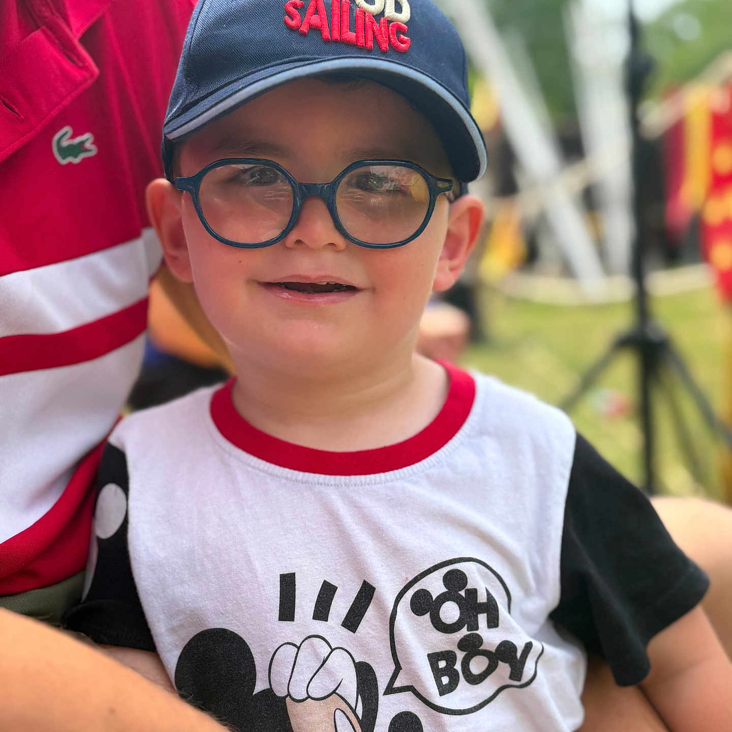 Lukas a rejoint le concours — aidez-le/la à gagner de superbes lots ! blurred_background, boy, cap, casual_clothing, child, daylight, flag, fun, glasses, happy, headwear, mickey_mouse, outdoor, person, portrait, red_shirt, sitting, smile, summer, tshirt