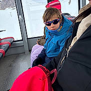 Enzo a rejoint le concours — aidez-le/la à gagner de superbes lots ! child, kid, sunglasses, blue_jacket, scarf, gloves, backpack, snow, mountains, gondola, cabin, window, seat, adult, winter_clothing, portrait, travel, reflection, ski_trip, pink_bag